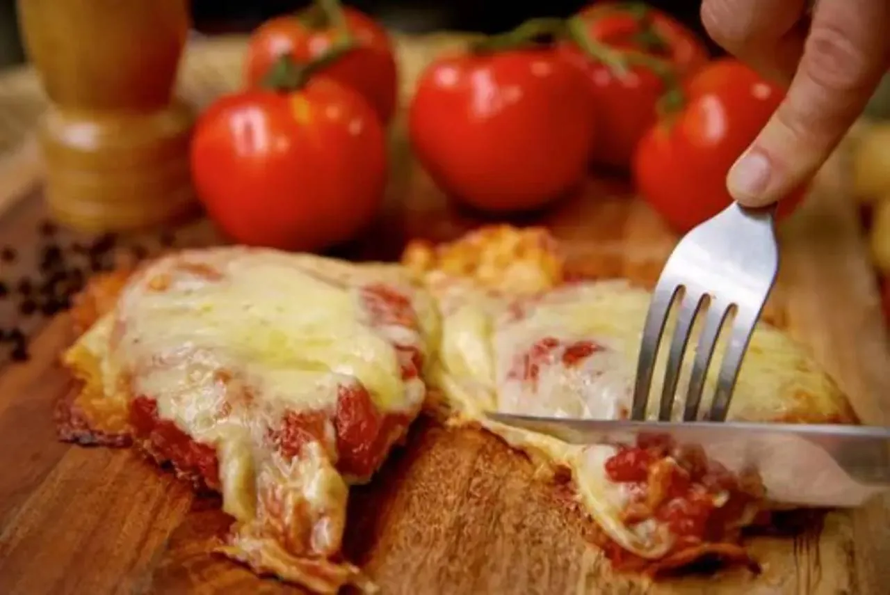 Filé de frango empanado à parmegiana com molho de tomate e queijo derretido, sendo cortado com garfo e faca sobre uma tábua de madeira, com tomates ao fundo.